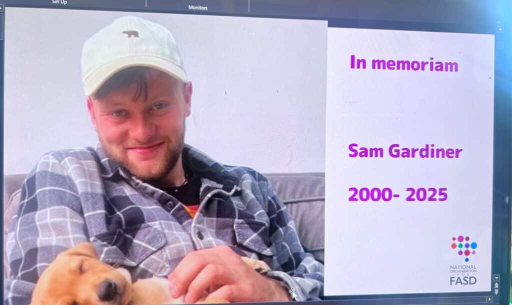 A photo of a presentation slide with a photo of Sam Gardiner, in a white cap and plaid shirt cuddling a tan dog. text says in memorium sam gardiner 2000-2025