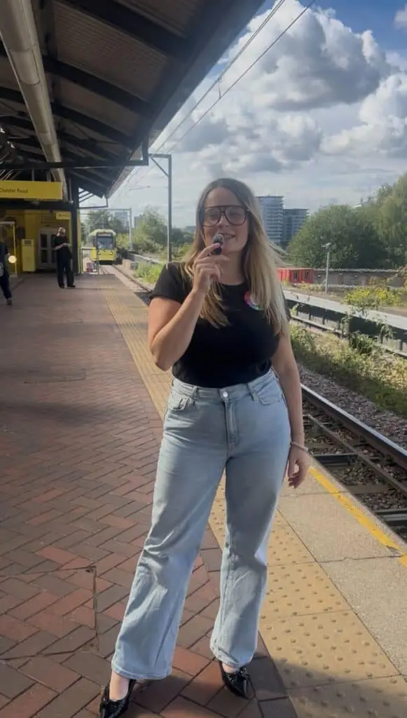 Alice Sewell holding a microphone standing by a tram track wearing an "Ask Me About FASD" badge